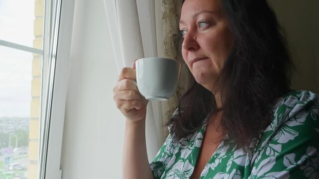 Woman in green floral shirt holds white mug while looking out window, capturing peaceful morning routine, relaxation, contemplation by natural light indoors.