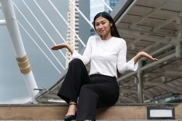 Smiling young Asian woman sitting on city stairs shrugging shoulders with open hands gesturing unsure or presenting isolated on modern urban architecture background with copy space. © kaew6566