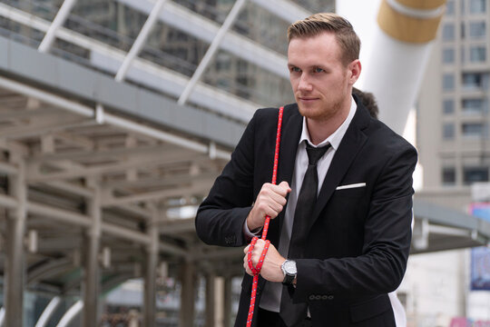 Determined caucasian businessman in black suit pulling red rope in tug of war challenge outdoors in modern city. Concept of corporate competition, business struggle, strength and team effort.