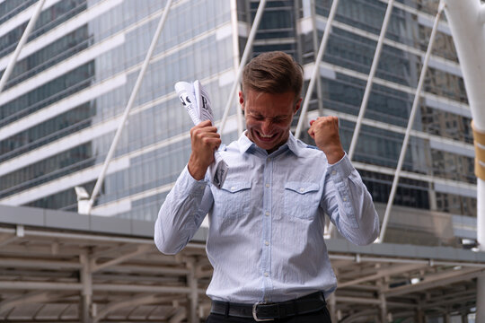 Excited caucasian businessman standing outdoors in modern city holding newspaper raising fists celebrating victory and career success. Concept of good news and winning deal.