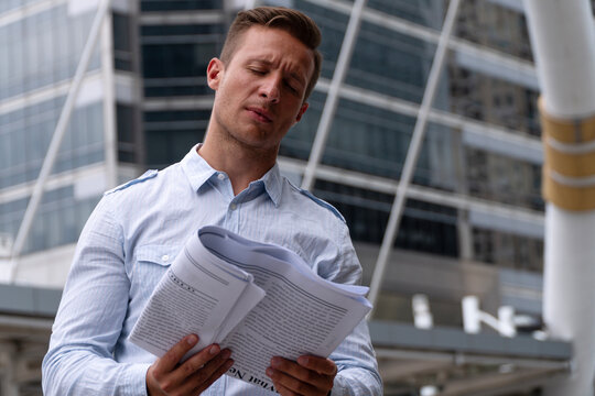 Stressed caucasian businessman standing outdoors in modern city holding paperwork closing eyes feeling tired and exhausted. Concept of corporate overwork and business problems.