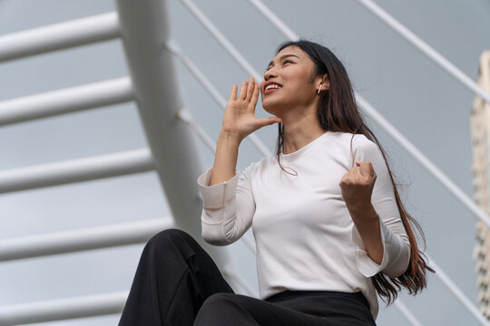 Excited young Asian woman sitting on city stairs putting hand to mouth shouting cheering and raising fist celebrating victory on modern urban architecture background with copy space.