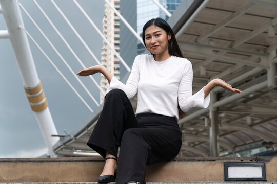 Smiling young Asian woman sitting on city stairs shrugging shoulders with open hands gesturing unsure or presenting isolated on modern urban architecture background with copy space.