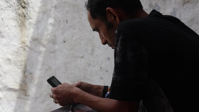  Indian man standing beside a worn wall, looking at his mobile phone with a thoughtful expression, reflecting on life, personal struggles, stress, and everyday concerns.