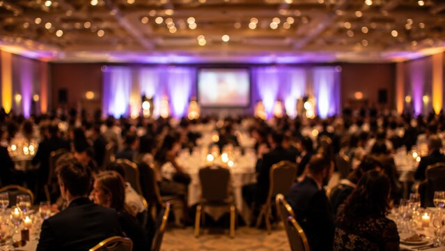 Large audience attends a formal gala dinner in a banquet hall. Guests sit at round tables while facing a stage with a projection screen under soft ambient purple lighting.
