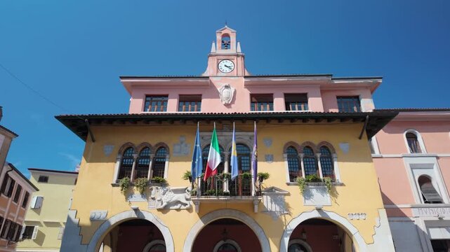 Historic Italian town hall of Muggia-Milje with yellow and pink facades, clock tower, arched windows, balconies, Italian, european and local flags, and ornate details beneath a clear blue sky.