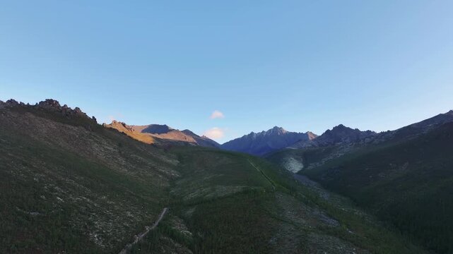 A wide aerial view of a mountainous valley with green forested slopes and rocky ridges under a clear blue sky. A single small cloud drifts slowly across the upper center during late afternoon.