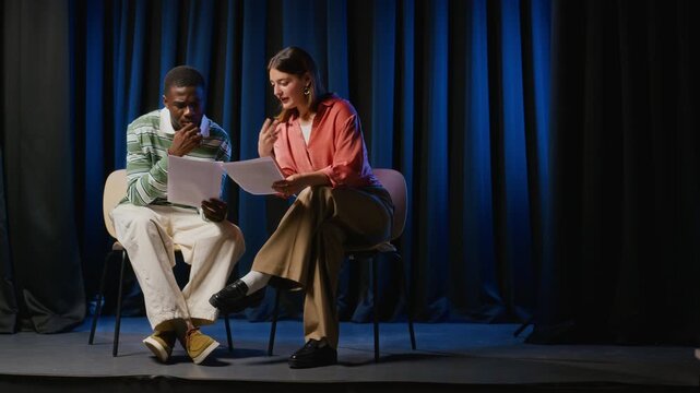 Young adult African American man and Caucasian woman reading theater scripts on stage during rehearsal. Ideal for promoting drama school education and university performing arts programs