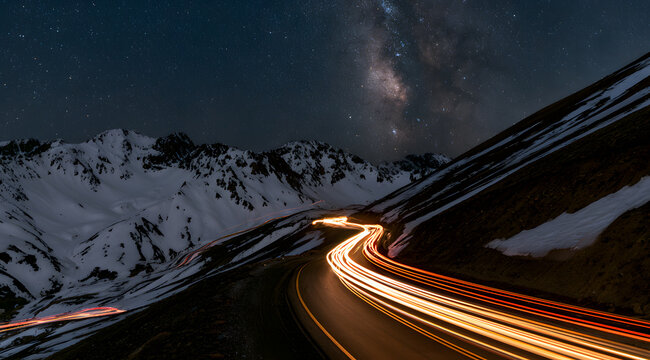 A sky ablaze with stars over a snowy mountain road, illuminated by the vibrant trails of passing cars.