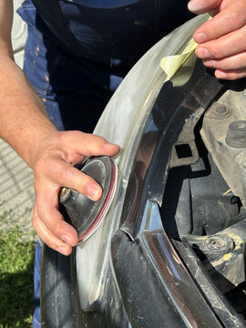 Close-up of a craftsman's hands sanding a plastic car headlight in Gomel. Masking tape protects edges. Active preparation of equipment for the autumn-winter season, August 28, 2026.