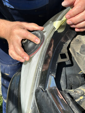 Close-up of a craftsman's hands sanding a plastic car headlight in Gomel. Masking tape protects edges. Active preparation of equipment for the autumn-winter season, August 28, 2026.