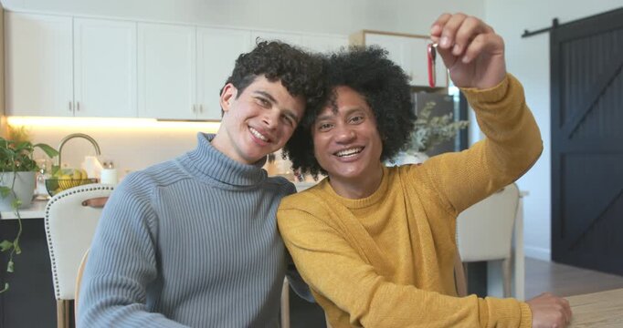 Male partners sitting close at home kitchen table, right-side man lifting red key fob celebrating