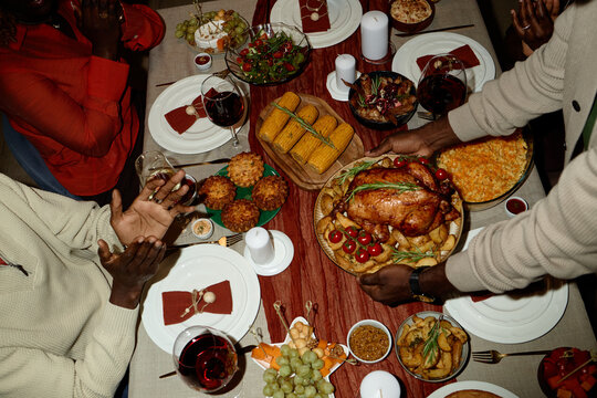 African American young adults sharing Thanksgiving dinner at home, hands serving roasted turkey. High angle view of festive holiday meal, ideal for seasonal family marketing campaigns