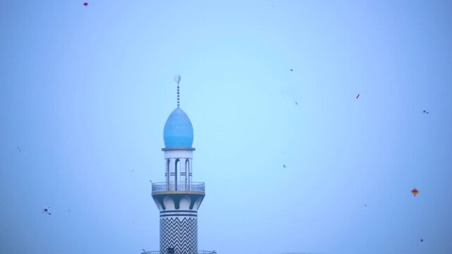 Mosque minaret rises beneath a pale sky with scattered kites drifting overhead.