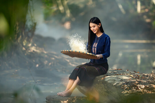 "The beauty of rural Thailand: Witness a young woman in handwoven indigo textiles expertly winnowing rice, capturing the heart of traditional Sakon Nakhon life. Ideal for banner and advertising use."