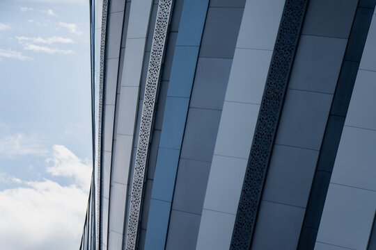 Angled metal cladding panels catch the light along a modern building facade, their perforated strips cutting sharp diagonal lines against a bright sky.