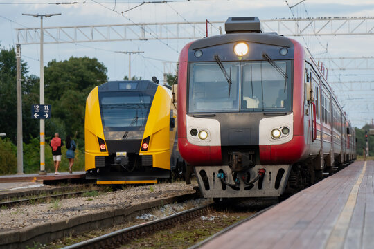 Headlights glow against overcast station light as an older red train arrives beside a waiting yellow regional unit, two generations of fleet at one platform.