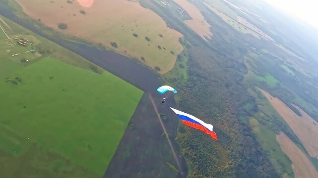 An aerial view of a parachutist with a large Russian flag, descending under a canopy over agricultural fields and a forested area.
