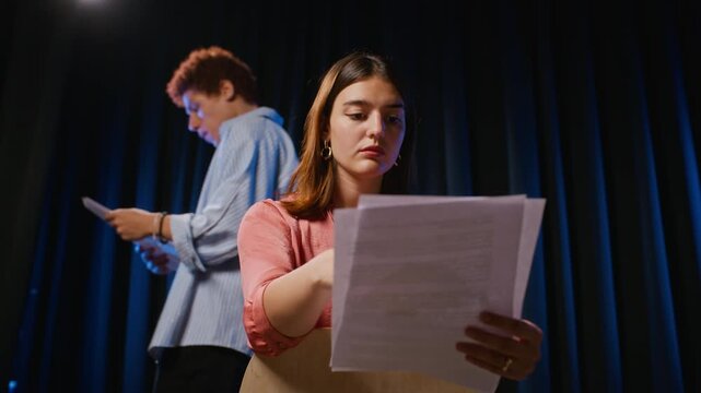 Caucasian young adult woman reading script during drama rehearsal on stage with actor in background. Footage suitable for performing arts education, acting classes, casting ads