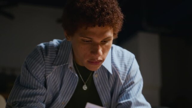 Young African American male student reading script and making notes with pen, camera tilting up to focused face. Useful for theater education, acting academy promo, performing arts casting videos