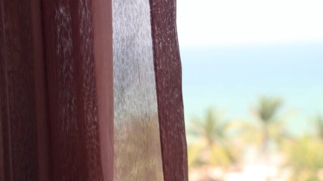 Soft transparent curtains gently blowing before a window with a blurred view of the tropical sea beach with palm trees