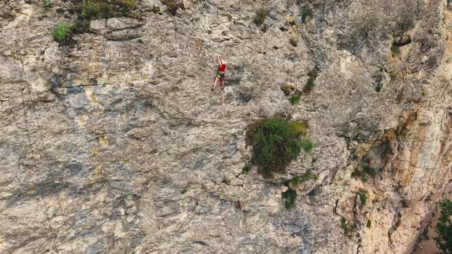 Climber in red ascends a rocky cliff. They focus intently on handholds carefully. The steep rock face has sparse vegetation. Lush green trees cover the valley below. The cliff slopes downward steeply