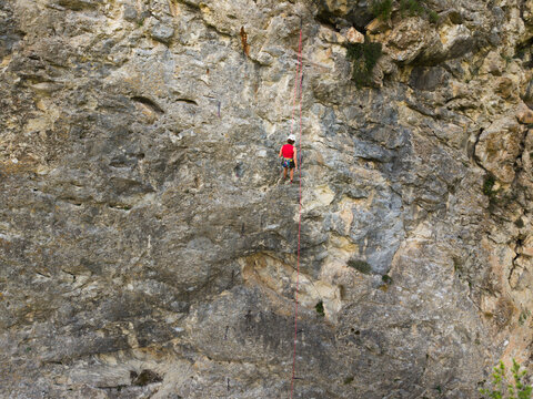 Gray rock face shows rough texture and weathered surface. Red climbing harness secured on stone. Attached rope runs vertically down the wall. Small green vegetation patches appear at the bottom