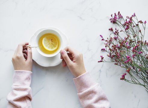 Top view of woman holding a cup of hot lemon tea beside dried purple flowers on a white marble background.