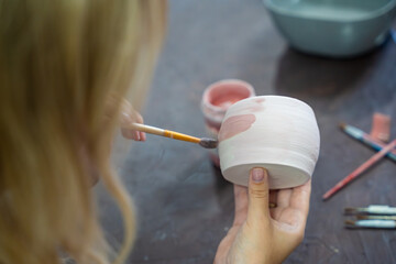 Woman painting pink glaze on handmade ceramic mug with brush in pottery workshop. Pottery decoration represents creativity and learning a craft as a hobby © dtatiana