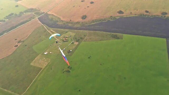 An aerial view of two parachutists over fields and an airfield. One athlete is descending with a large Russian tricolor.
