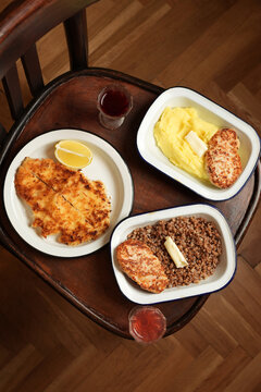 High angle flat lay of schnitzel, chicken cutlets with mashed potatoes and buckwheat porridge served in enamel dishes with shot glasses on a wooden chair