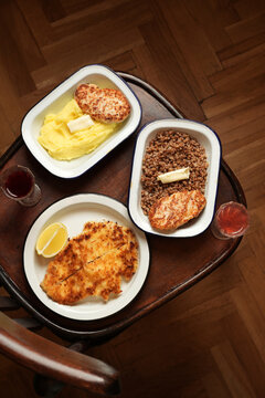 High angle flat lay of schnitzel, chicken cutlets with mashed potatoes and buckwheat porridge served in enamel dishes with shot glasses on a wooden chair