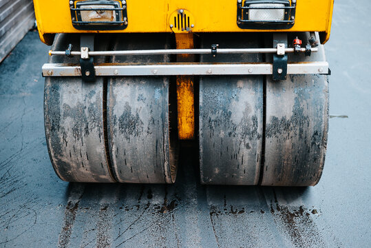 A heavy road roller is seen compacting asphalt on a construction site. The large cylindrical tires show signs of wear and are set against a wet surface reflecting the surroundings