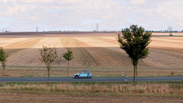 Vintage retro small car drives along rural road beside harvested fields under cloudy sky. Scenic countryside scene combines classic motoring, agriculture, travel, nostalgia, and calm open landscape