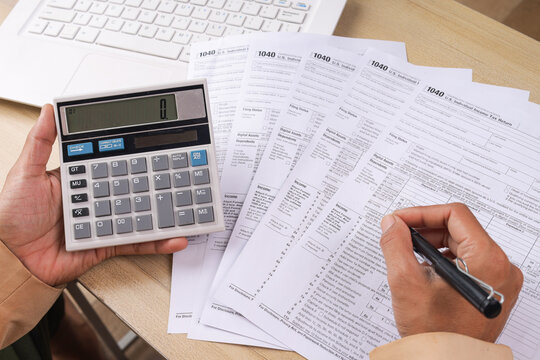 Hand Holding Pen to Calculate 1040 Tax Return Beside Laptop