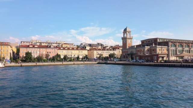 Historic waterfront lined with colorful buildings of Trieste, a clock tower, and a promenade, viewed from approaching boat.