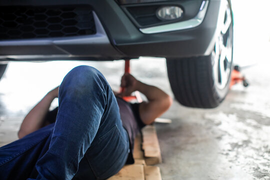 Selective focus to leg of mechanic lying on the ground under a lifted car performing undercarriage maintenance and repairs in a garage shop.