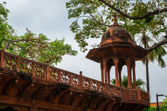 Red sandstone pavilion and intricate carved balcony surrounded by green tree branches against overcast sky at Taj Mahal, Agra, Uttar Pradesh, India.