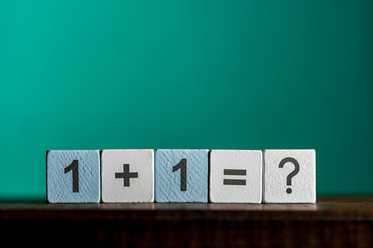 Simple math equation on wooden blocks with question mark, closeup. White and light blue wooden cubes showing the mathematical equation one plus one equals question mark stand against green background