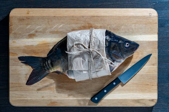 Fresh carp fish wrapped in craft paper with knife, closeup. Top-down view of fresh raw carp fish wrapped in brown parchment paper and tied with twine next to kitchen knife on wooden cutting board