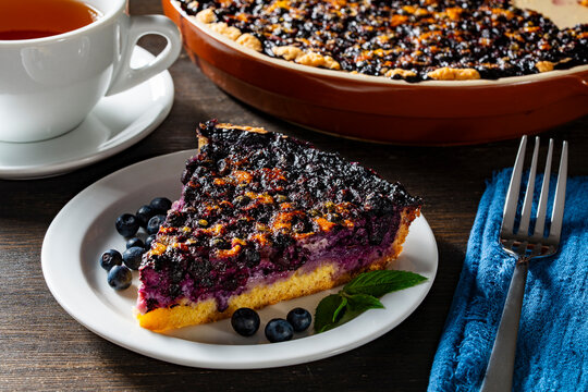 Slice of homemade blueberry pie with fresh berries and tea. Slice of delicious blueberry pie served on white plate with fresh berries and mint next to cup of tea and baking dish on wooden table