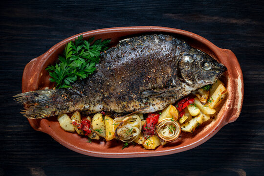 Baked carp fish with potatoes and vegetables, close up. Whole baked carp fish seasoned with spices, served with roasted potatoes, onions, and tomatoes in rustic clay baking pan on dark wooden table