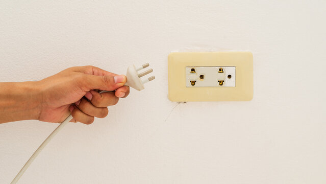 Hand holding three pin power plug near wall socket outlet