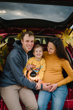 Happy family sitting in car trunk decorated with autumn leaves and holding lantern. Pregnant mom, smiling dad, and son together outdoors during a road trip.