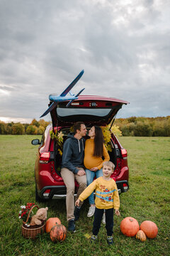 Toy airplane flying over family near car trunk decorated with autumn leaves and pumpkins. Happy mom, smiling dad, and son outdoors during a road trip, with basket on the grass. Kid playing toy plane.