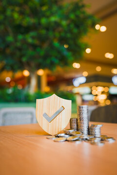 Financial security and deposit insurance concept with wooden shield with checkmark beside stacks of coins on table against warm bokeh background for guaranteed savings and capital protection.