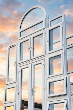 Great composition of new white PVC windows and glass doors stacked outdoors against a beautiful orange sunset sky. Concept of home renovation, construction, energy efficiency, and house.