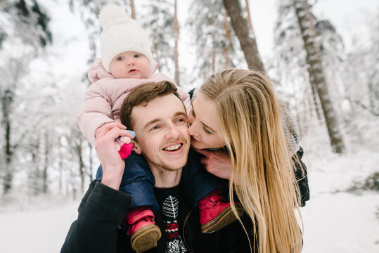 Happy parents in snowy forest, mother tenderly kissing smiling father who carries their little daughter on his shoulders. Family love, winter mood and parental happiness. Closeup.