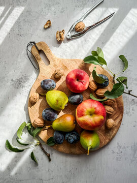 Reifes Obst auf einem runden Holz Schneidebrett und ein Nussknacker auf einem grauen Tisch im Sonnenlicht. Vertikal, Herbst.