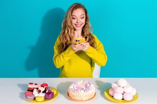 Young woman smiling at smartphone with colorful desserts on table in bright blue studio